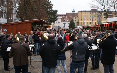 Das Bild zeigt eine Blasmusikkapelle sowie mehrer Buden und Besucher des adventlichen Marktes.